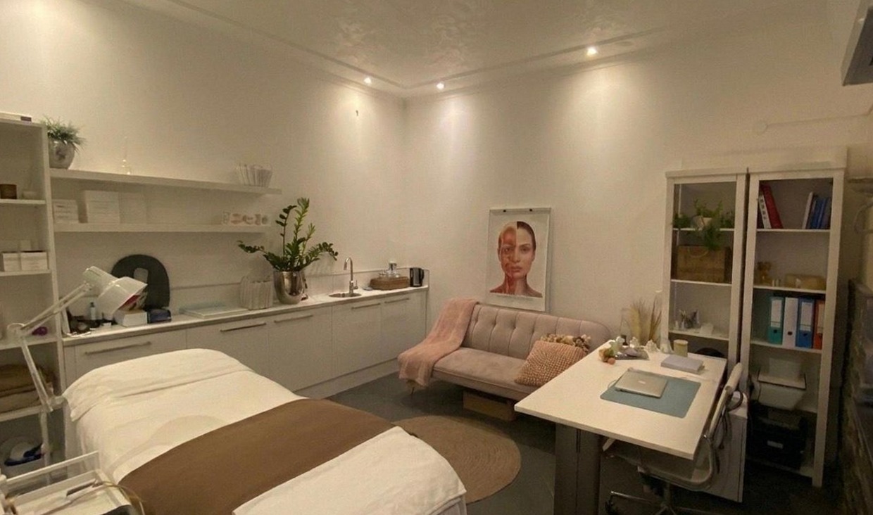 De behandelkamer van Bruna Marins Institute: behandelbank met taupe loper, witte kasten, een roze bank en een anatomieposter aan de muur